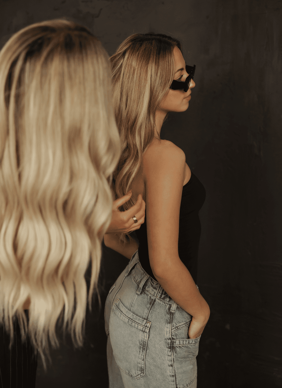 Two women with long blonde hair; one adjusting the other's hair against a dark background.