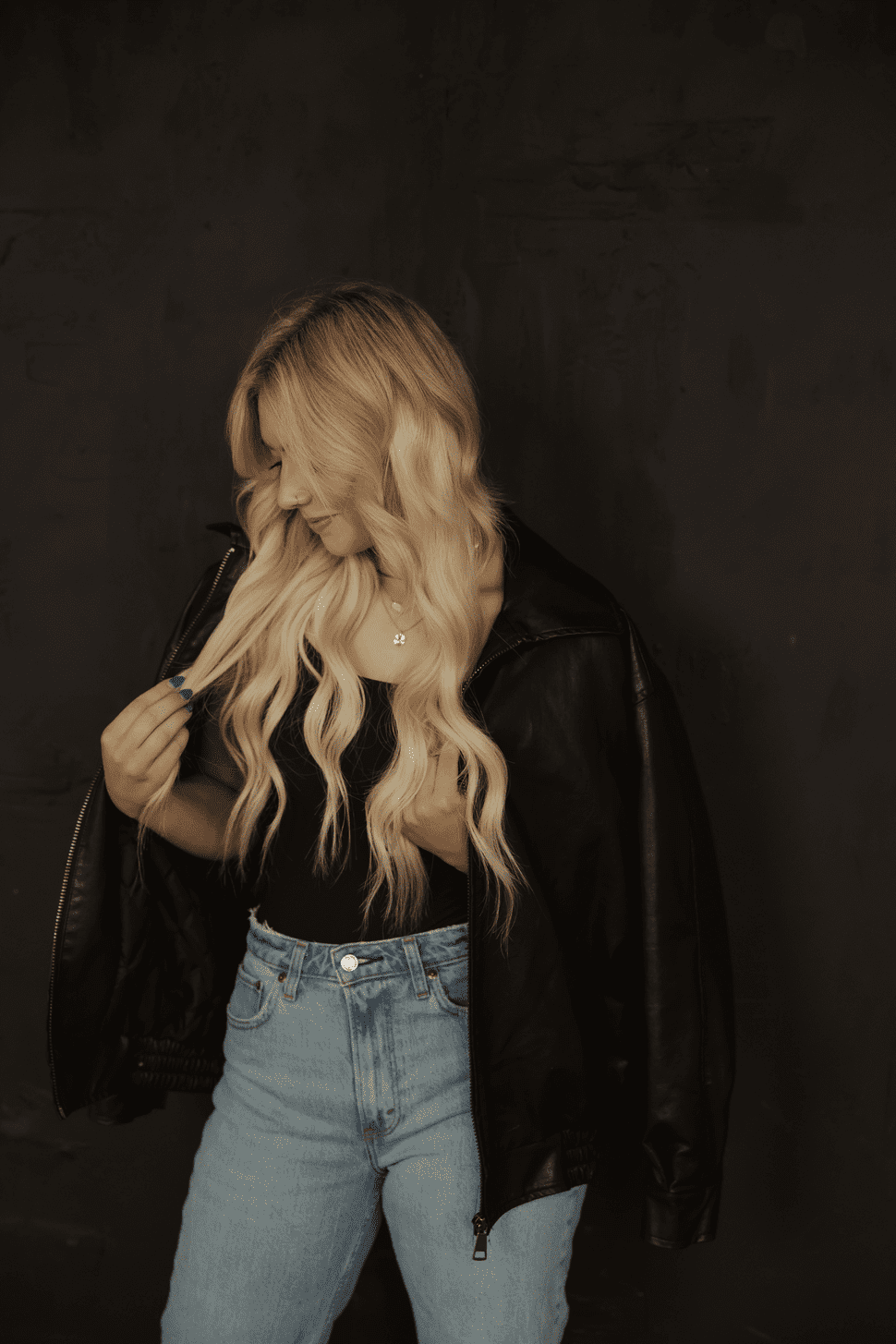 Blonde woman in black leather jacket and jeans, looking down, against a dark background.